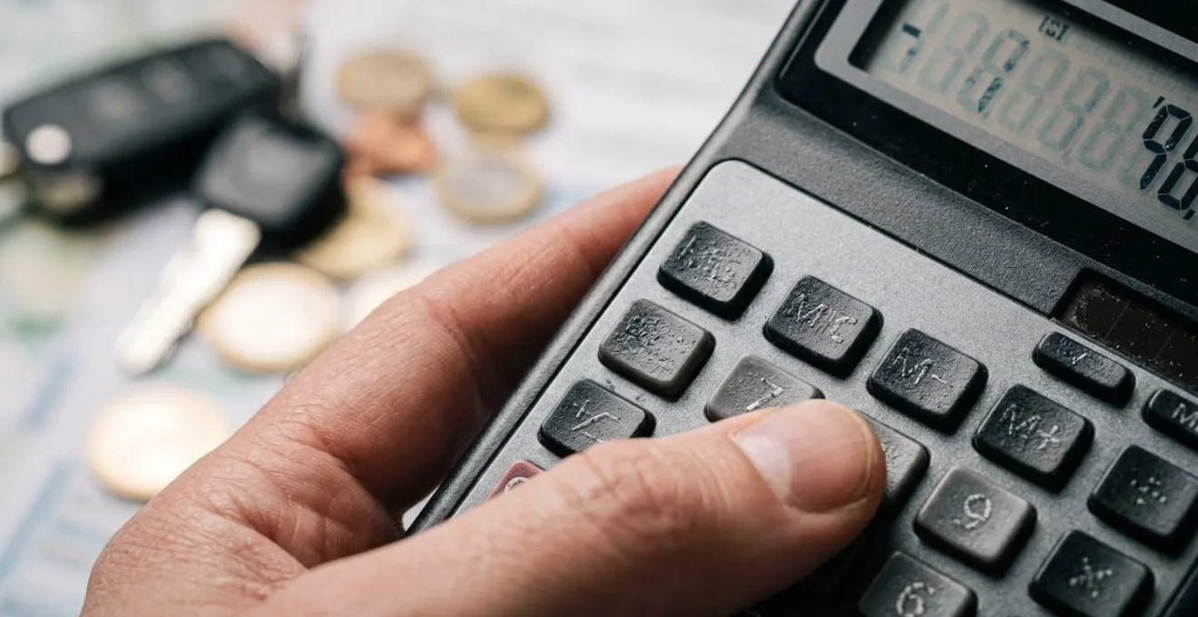 Main tenant une calculatrice avec des pièces automobiles et papiers administratifs flous en arrière-plan